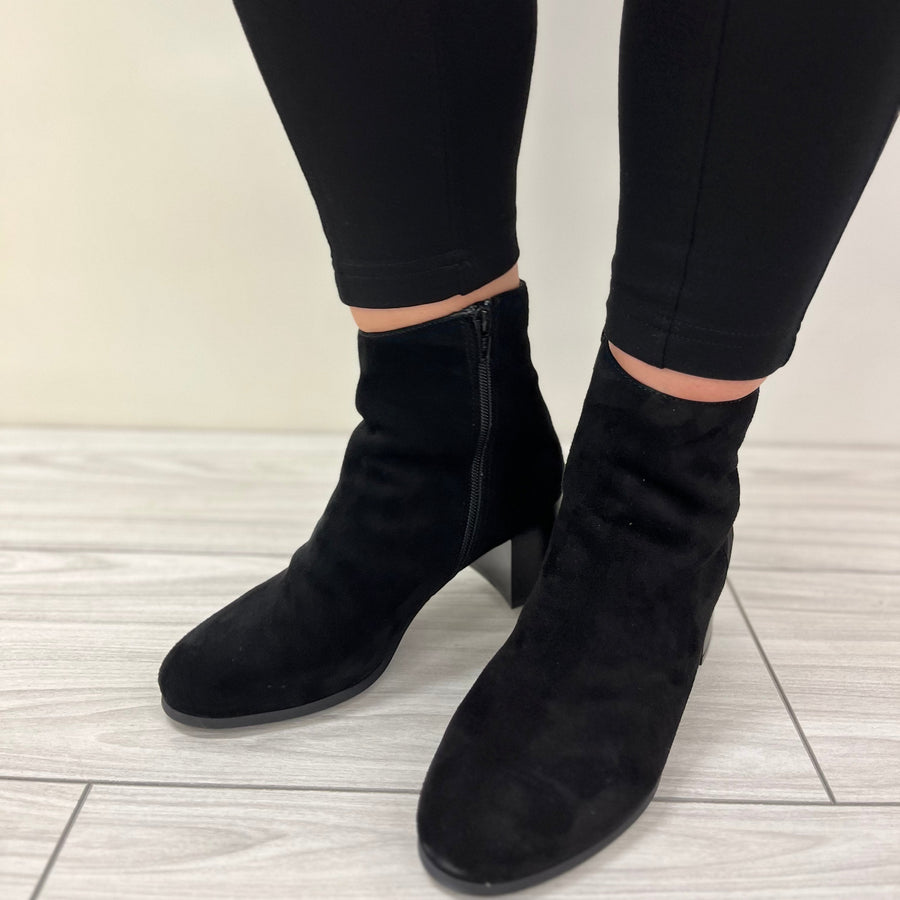 Black ankle boots worn with black pants on a light wooden floor.