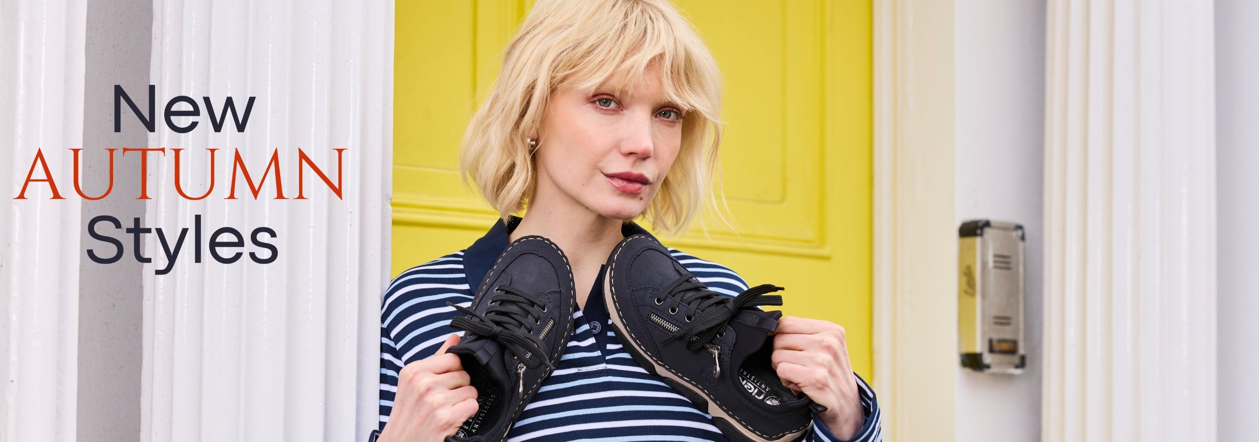 A woman with blonde hair wearing a navy and white striped shirt holds up a pair of black lace-up shoes in front of a bright yellow door. Text on the left reads 'New AUTUMN Styles'.